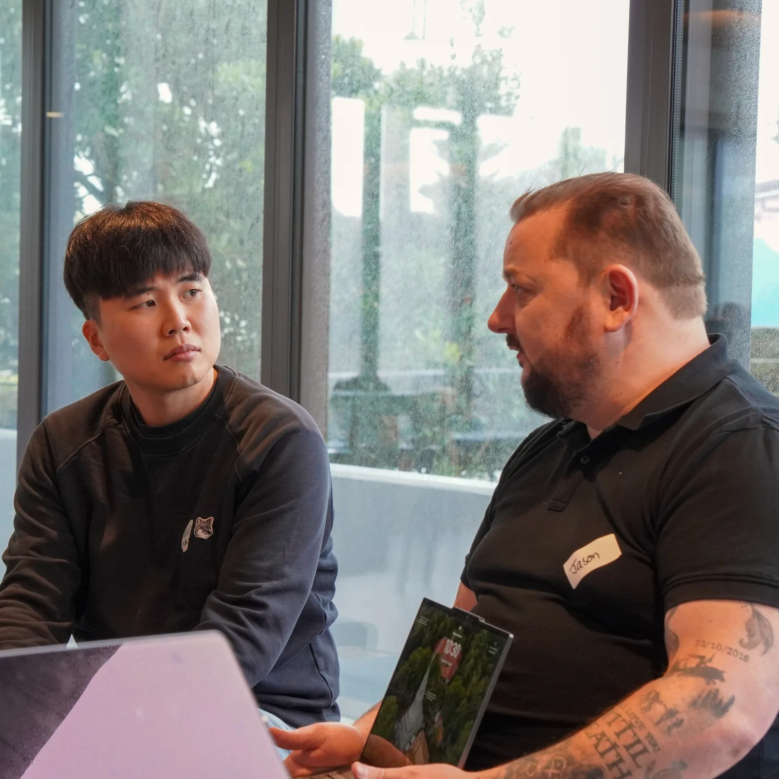 Two people talk through a project at their laptops beside a rain-flecked window during a For Good build session.