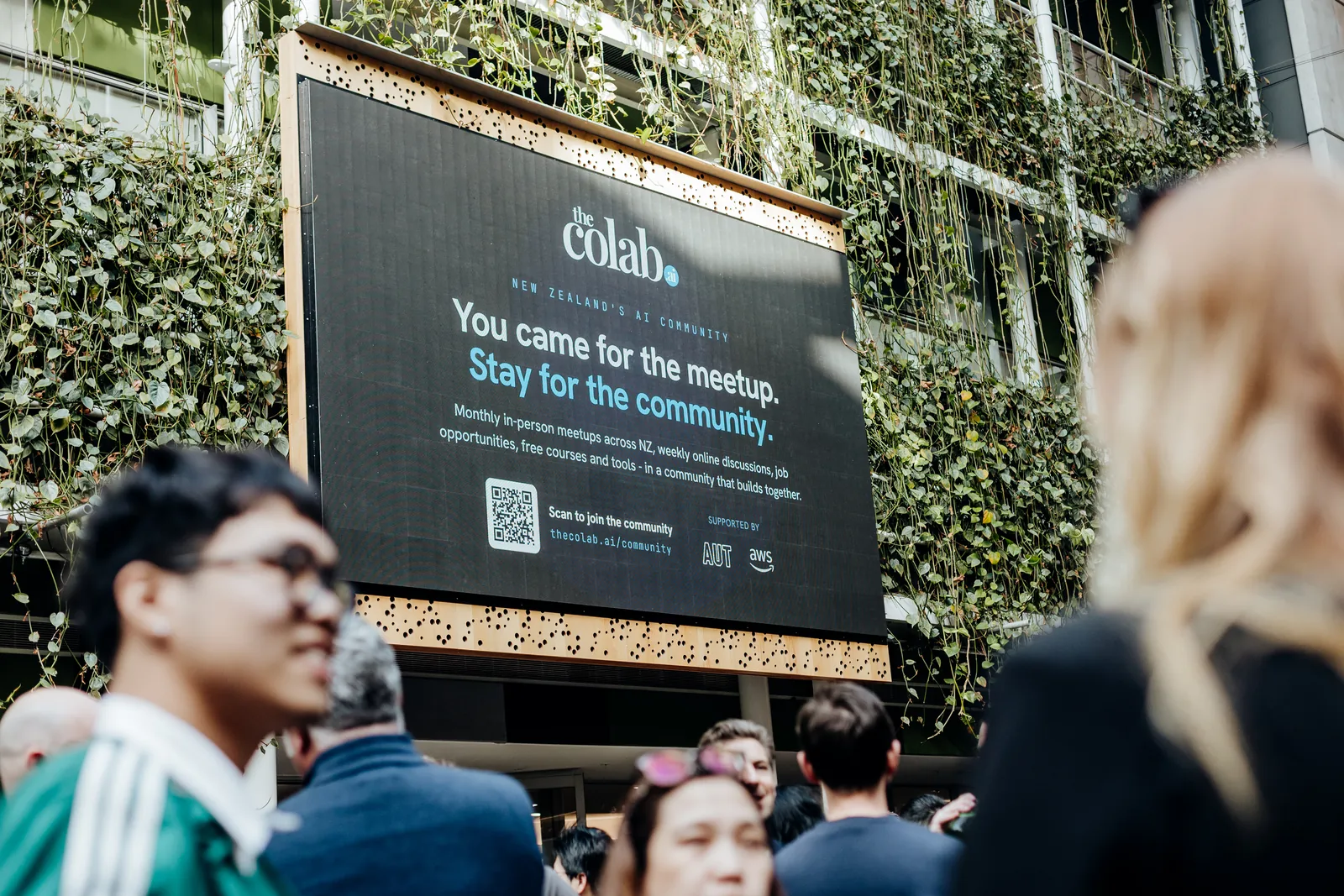 Meetup guests gather under the big screen in the AUT atrium, which reads 'theColab: You came for the meetup. Stay for the community.' with a QR code to join.