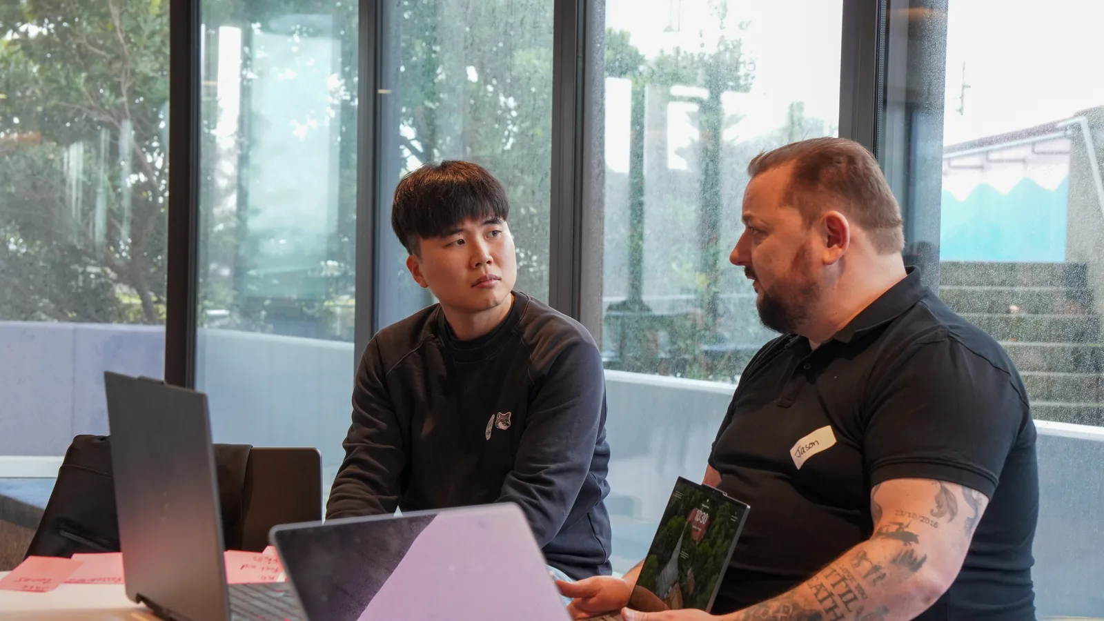 Two participants at a laptop-covered table talking through an idea, one holding a tablet, rain-streaked windows behind them.