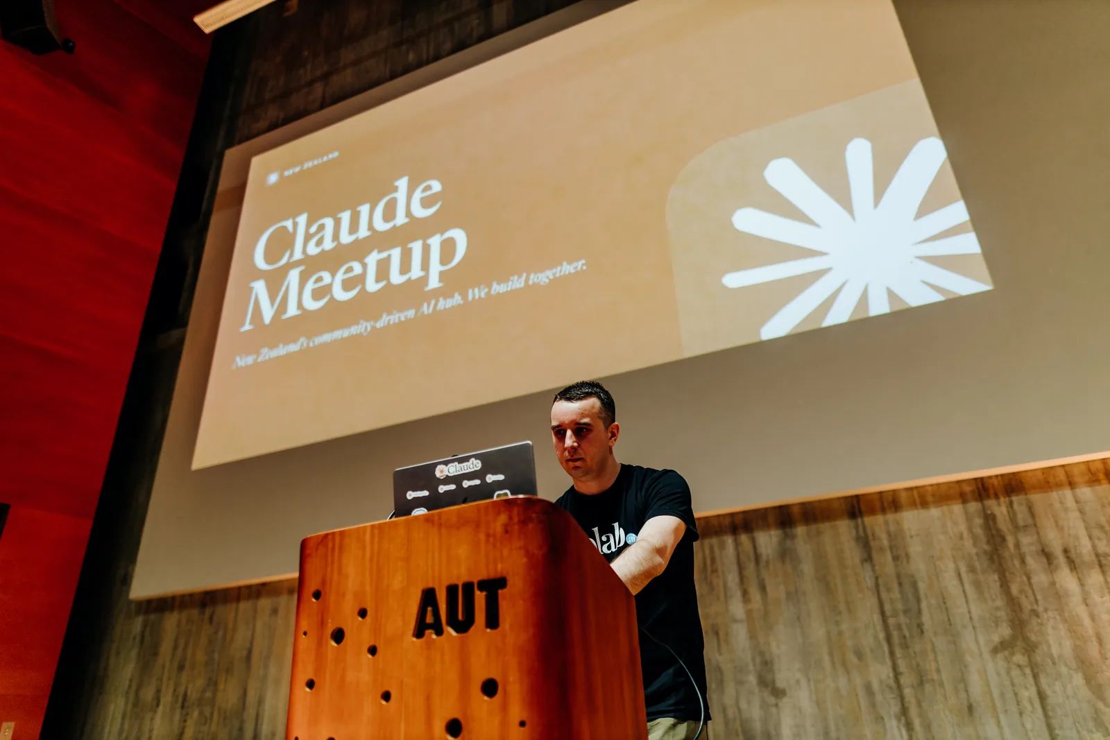 Adam Holt, Colab co-founder, at the AUT lectern with his laptop, under a big 'Claude Meetup' title slide.
