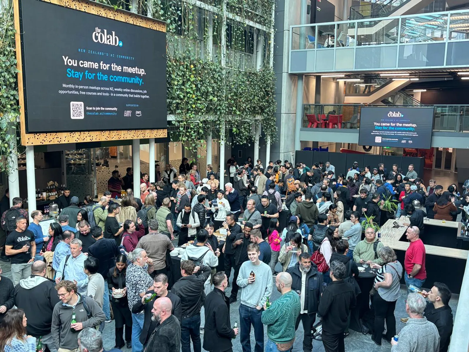 A big crowd fills the AUT atrium at a thecolab.ai community meetup, chatting over drinks under the 'You came for the meetup. Stay for the community.' screen.